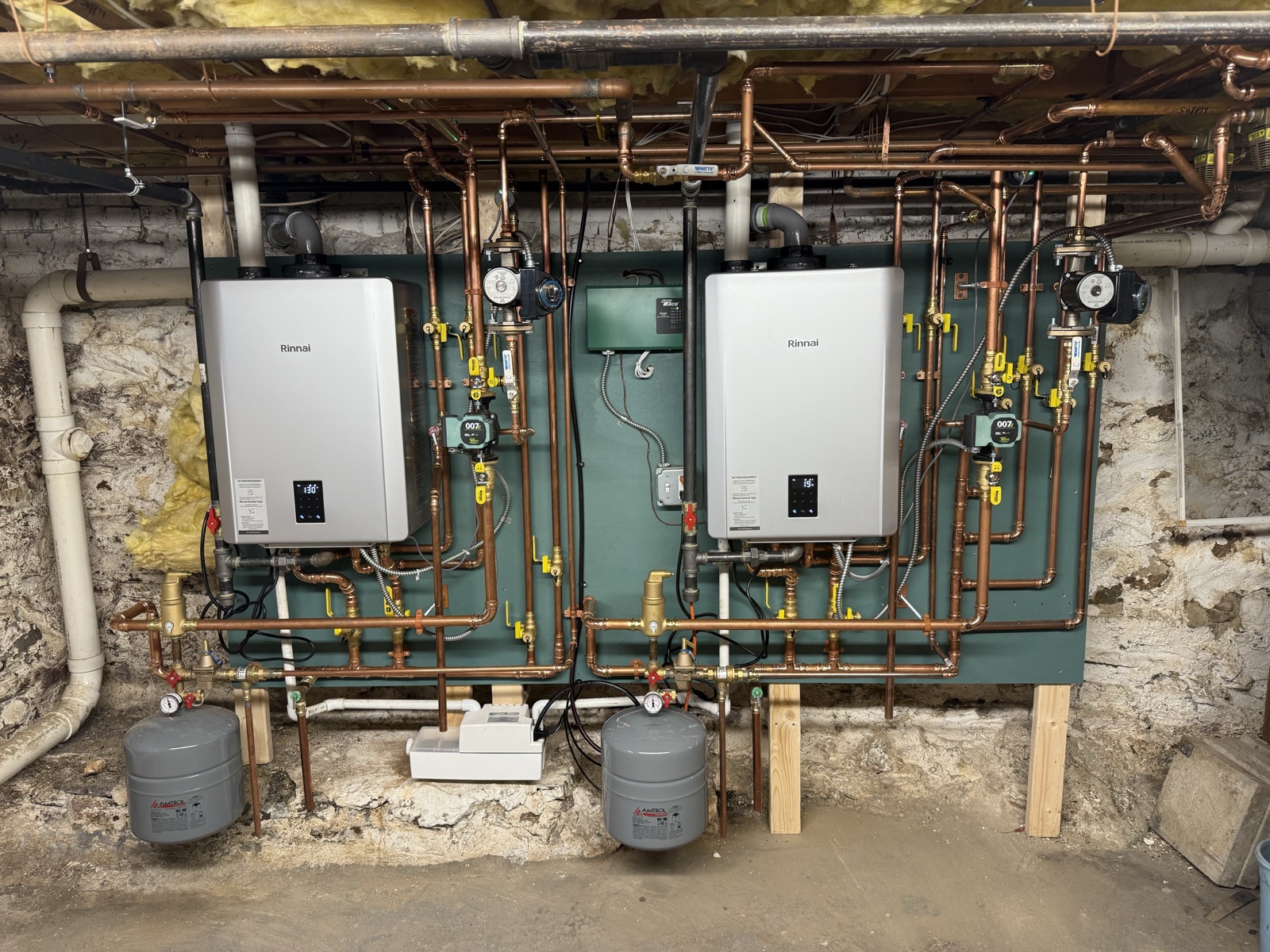 Two Rinnai tankless water heaters on a painted backer board mounted against an old stone foundation